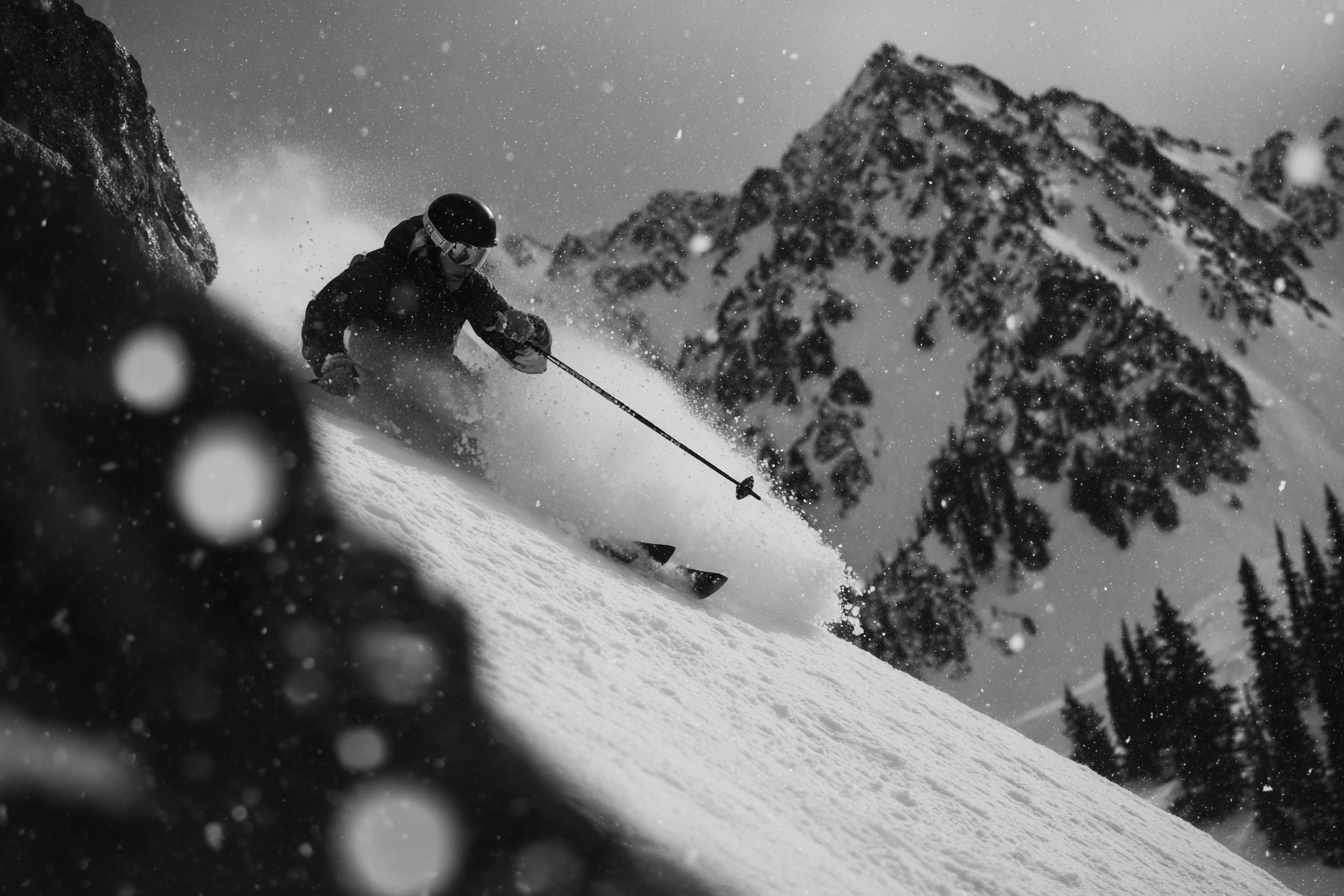 Skiër maakt een bocht door diepe poedersneeuw met besneeuwde bergtoppen en dennen op de achtergrond tijdens sneeuwval