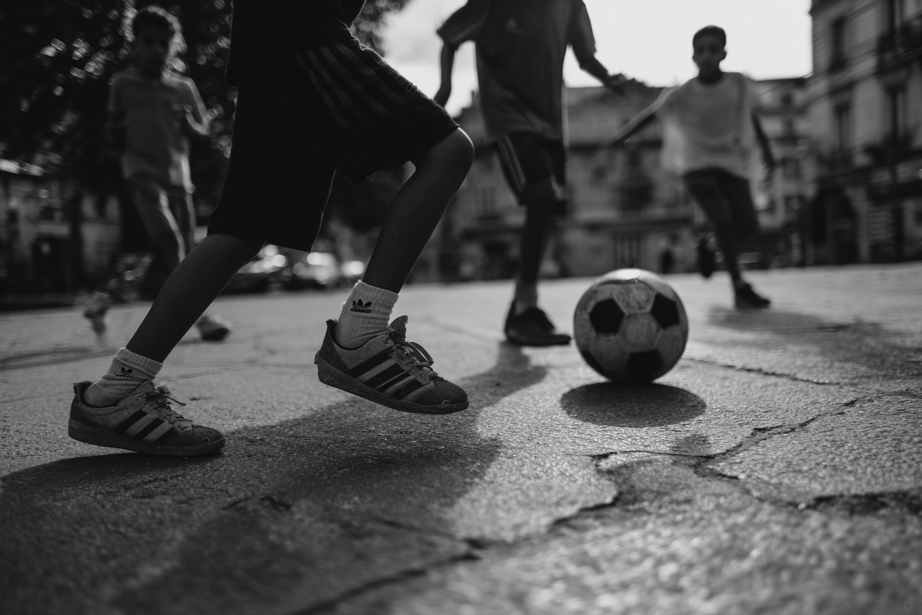 Kinderen spelen voetbal op een verweerd stadsplein, met een bal en sneakers op de voorgrond