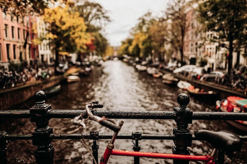 Fiets tegen een brugleuning boven een Amsterdamse gracht met herfstbomen, grachtenpanden en afgemeerde boten
