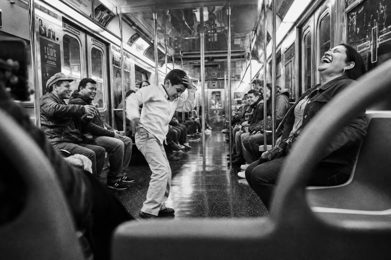 Jongen danst in het gangpad van een metrostel terwijl medereizigers lachend toekijken
