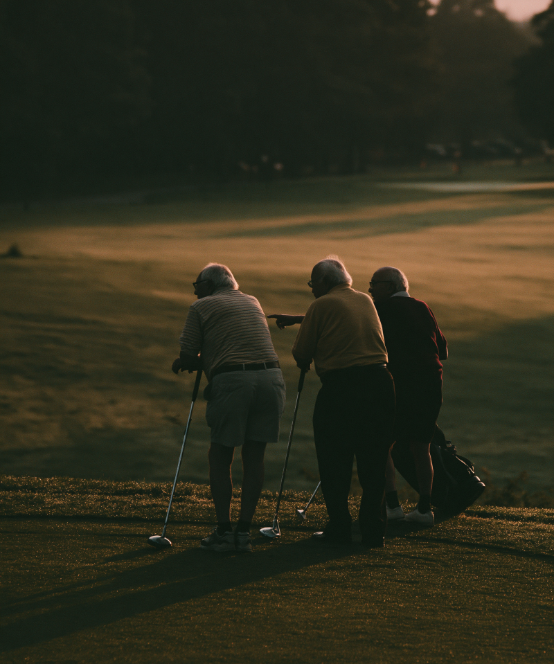 Drie oudere golfers overleggen op een golfbaan in warm avondlicht, een van hen wijst naar de fairway