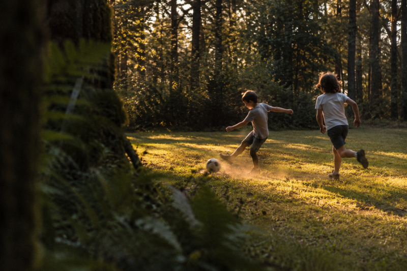 Twee kinderen spelen voetbal op een grasveld aan de bosrand in warm avondlicht