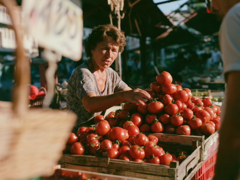 Vrouw achter een marktkraam schikt een stapel rijpe rode tomaten in houten kisten in warm ochtendlicht