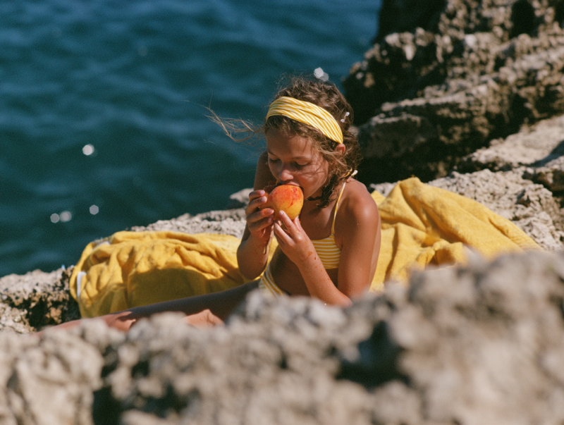 Jonge vrouw in gele bikini bijt in een perzik terwijl ze tussen rotsen aan het diepblauwe water zit