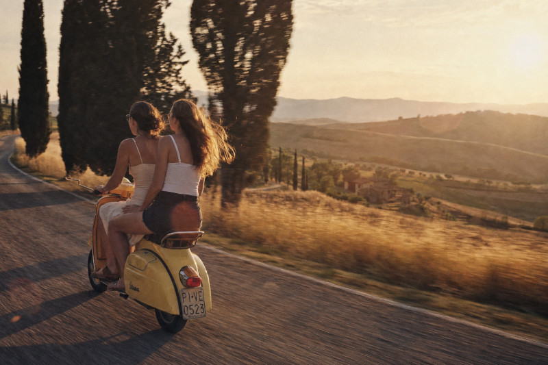 Twee vrouwen rijden op een gele scooter over een landweg door glooiende Toscaanse heuvels bij zonsondergang
