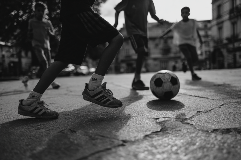 Kinderen spelen voetbal op een verweerd stadsplein, met een bal en sneakers op de voorgrond