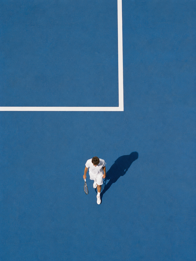 Tennisser in het wit met racket loopt over een blauwe baan, gezien van bovenaf met een lange schaduw en witte lijnen