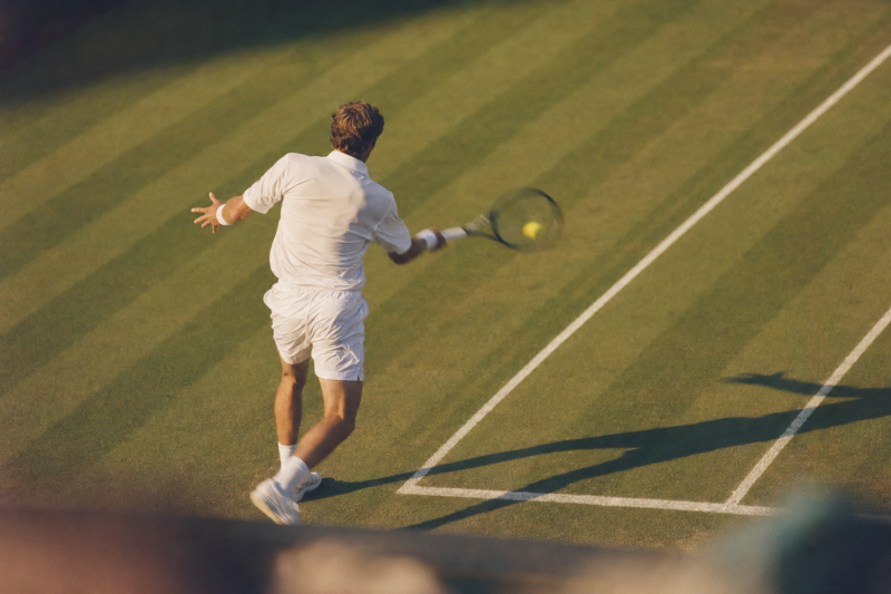 Tennisser in wit tenue slaat een forehand op een zonovergoten grasbaan