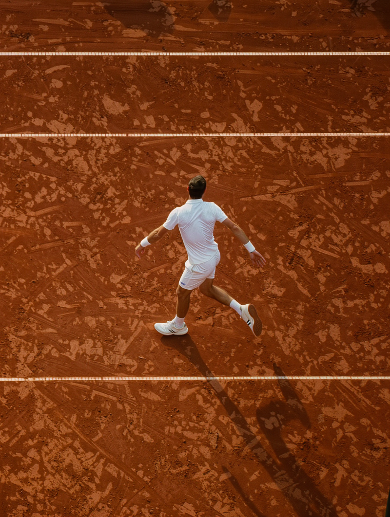 Tennisser in wit tenue rent over een roodbruine gravelbaan met witte lijnen van bovenaf gezien