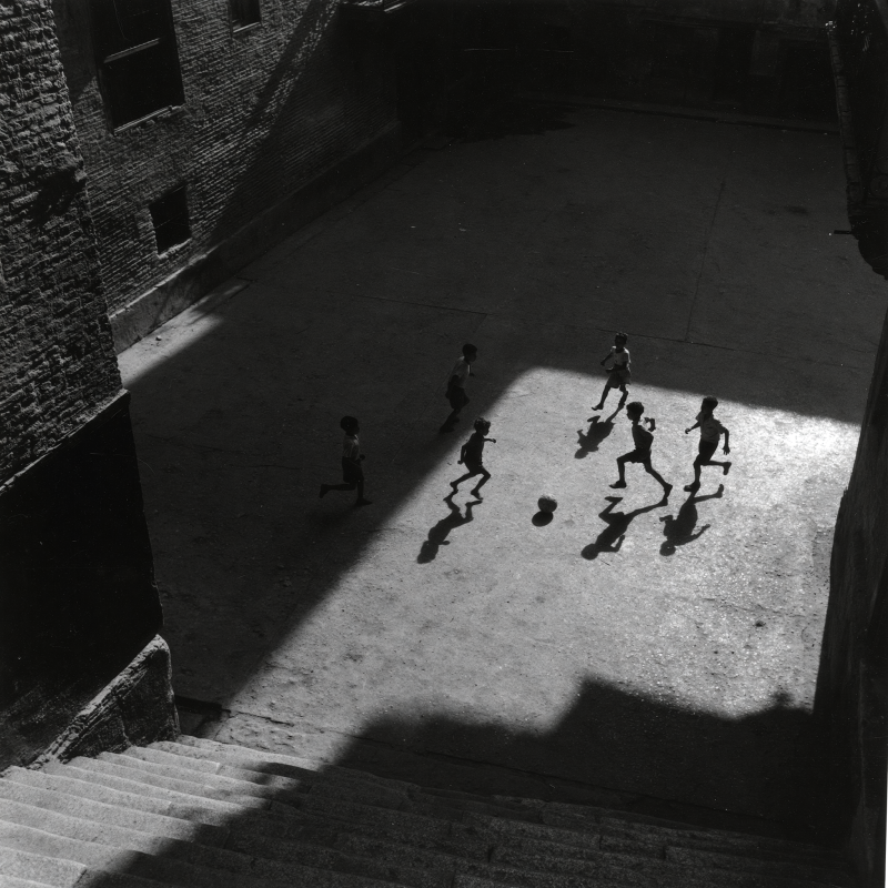 Groep kinderen speelt met een bal op een schaduwrijke binnenplaats waar een bundel zonlicht op het plaveisel valt