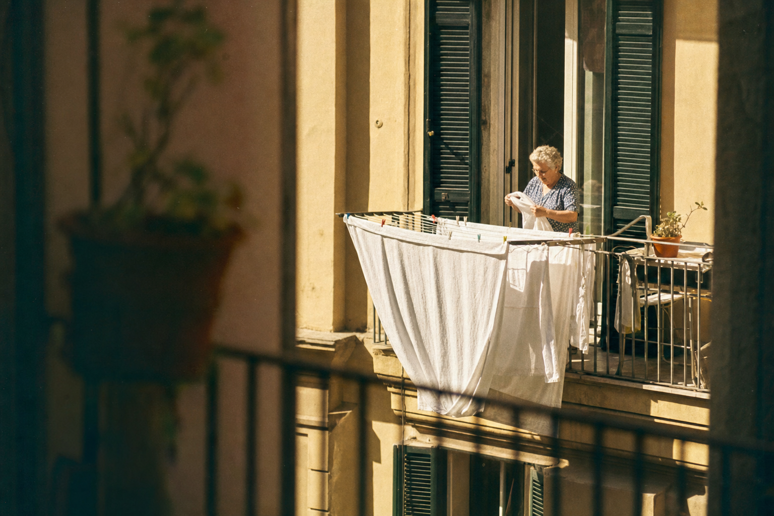 Oudere vrouw hangt witte was op aan een balkon van een geel gebouw in warm ochtendlicht, met potplanten en groene luiken